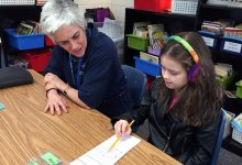 An young girl with rainbow-knitted headphones is sitting at a table with her female teacher. They're both looking at a long, thin sheet of paper, and the young girl is holding the tip of the pencil against the paper, pointing at something.